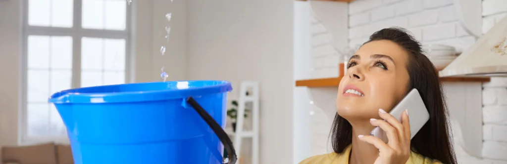 woman on phone looking up at leaking ceiling holding bucket